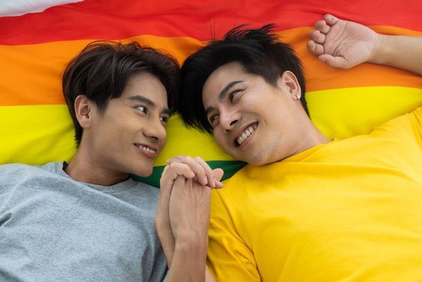 Aasialainen homopari pitää toisiaan kädestä sängyllä. Sateenkaaren lippu LGBT-merkkinä taustalla. Sukupuolten tasa-arvo ja oikea käsite, leikkisä ja romanttinen hetki. - Valokuva, kuva