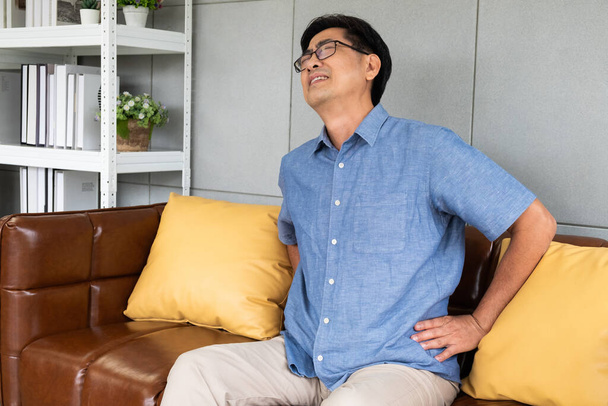 シニアアジア人男性の屋内ショットはソファのソファの上に座って、背中に触れ、背中の痛み、痛みの筋肉、骨やランバゴに苦しんでいます。高齢者の健康問題の概念. - 写真・画像