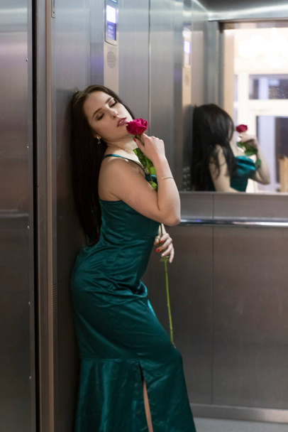 sexy mujer morena confiada en elegante vestido de color aguamarina con rosa roja en ascensor gris. espacio de copia - Foto, Imagen