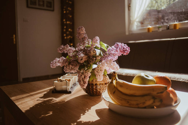 A selective focus shot of fresh fruits in the plate and lilac flowers in the vase in sunlight - 写真・画像