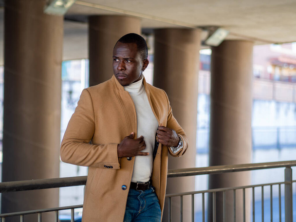 A shallow focus of a stylish African male wearing jeans with a white turtleneck and beige coat and leaning on a fence - Foto, Imagem