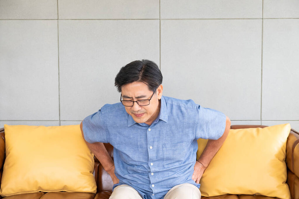 Vnitřní záběr starších asijských mužů sedí na pohovce, dotýkají se jeho zad a trpí bolestí zad, bolavými svaly, kostmi nebo bederní páteří. Starší koncepce zdravotního problému. - Fotografie, Obrázek