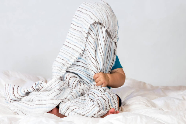 A baby sitting in a soft bed and hiding his face with a blanket - Zdjęcie, obraz