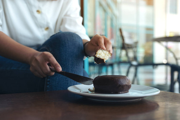 Detailní obrázek ženských rukou řezání kus čokoládové koblihy jíst nožem a vidličkou v bílém talíři - Fotografie, Obrázek