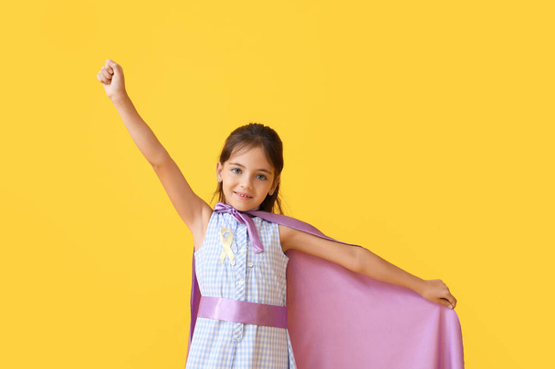 Little girl with golden ribbon wearing superhero costume on color background. Childhood cancer awareness concept - Photo, Image