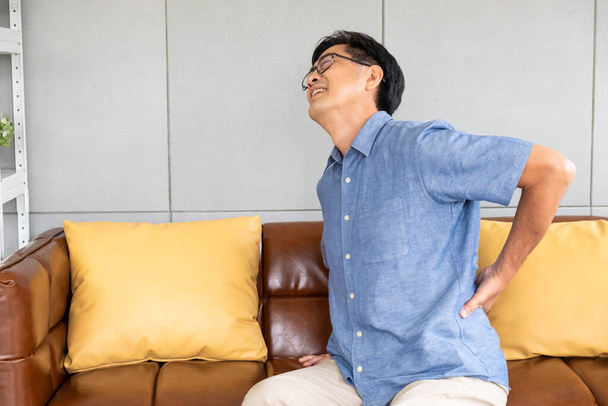 シニアアジア人男性の屋内ショットはソファのソファの上に座って、背中に触れ、背中の痛み、痛みの筋肉、骨やランバゴに苦しんでいます。高齢者の健康問題の概念. - 写真・画像