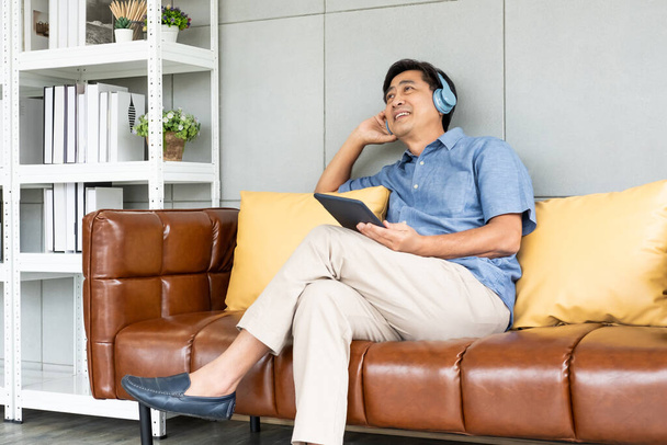 Выборочный фокус в лицо. Крытый снимок старших азиатских мужчин, сидящих на диване, слушая музыку из беспроводных наушников или смотреть онлайн фильм с цифрового планшета. Активное старшее поколение. - Фото, изображение