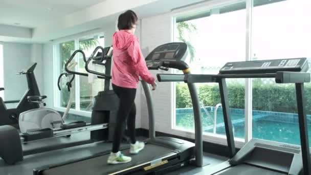 Gimnasio Mujer asiática en abrigo rosa empezar a caminar para calentar en la máquina de la cinta de correr, entrenamiento maratón en gimnasio de fitness - Imágenes, Vídeo