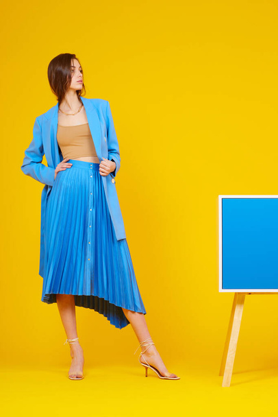High fashion portrait of young elegant woman in blue jacket and skirt. Yellow background, studio shot - 写真・画像