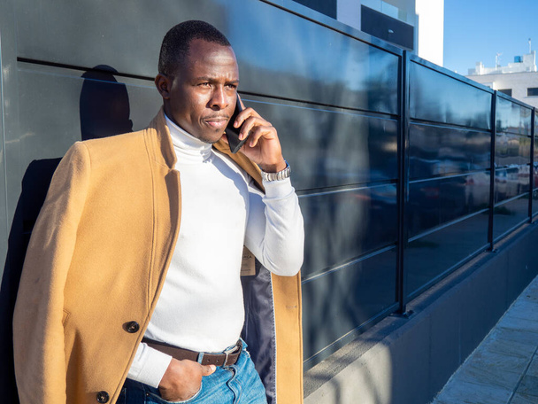 Egy elegáns, felnőtt afrikai férfi, aki a kerítésre támaszkodik, és telefonon beszél a napfény alatt. - Fotó, kép