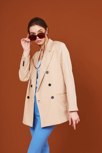 High fashion portrait of young elegant woman in beige jacket and jeans. Sunglasses, brown background, studio shot - Foto, afbeelding
