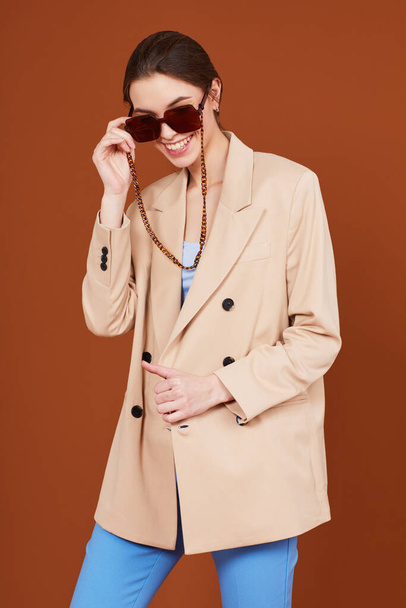 High fashion portrait of young elegant woman in beige jacket and jeans. Sunglasses, brown background, studio shot - Фото, зображення