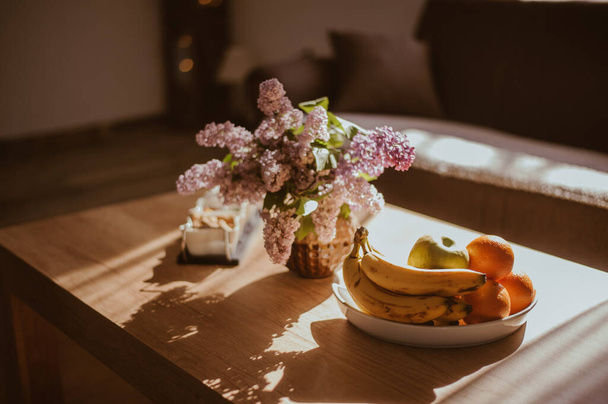 A selective focus shot of fresh fruits in the plate and lilac flowers in the vase in sunlight - Fotografie, Obrázek