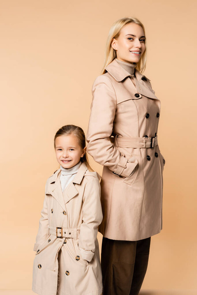 トレンチコートを着たスタイリッシュな母親と娘がベージュに隔離されたポケットに手を取りながら微笑む - 写真・画像