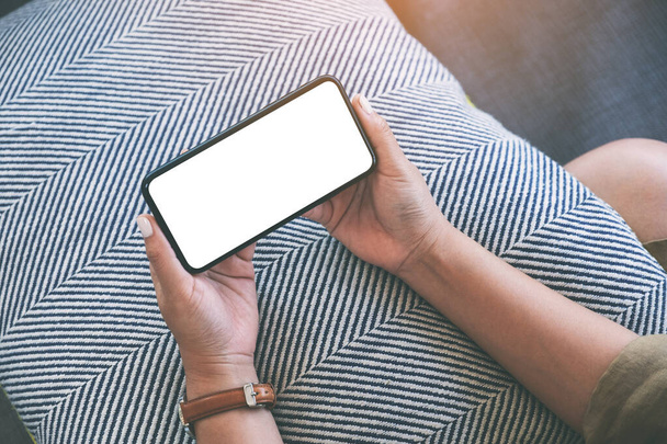 Mockup-Bild einer Frau, die schwarzes Handy mit leerem weißen Desktop-Bildschirm horizontal hält, während sie im Wohnzimmer sitzt und sich entspannt fühlt - Foto, Bild