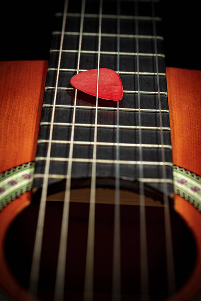 Extrême gros plan d'un pic de guitare rouge (plectrum) entre les cordes du manche d'une vieille guitare acoustique. Instrument à cordes. - Photo, image