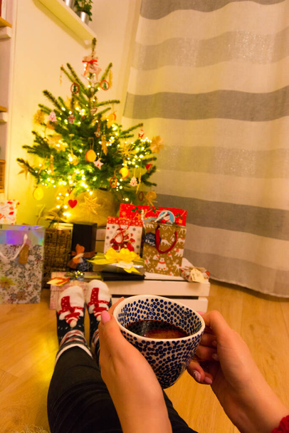 A vertical shot of female hands holding a cup of coffee next to a Christmas tree at home - Foto, imagen