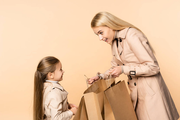 smiling mother holding paper bags near cheerful daughter isolated on beige - Fotografie, Obrázek