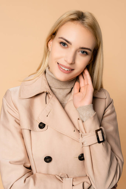 blonde woman in trench coat smiling and looking at camera isolated on beige - Photo, Image