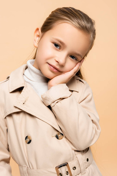 pleased child in trench coat looking at camera isolated on beige - Foto, Imagem