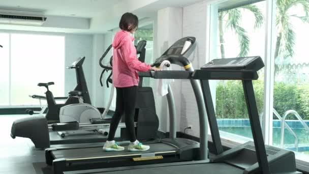 Fitness Asian woman in pink overcoat hang towel and push button setting to increse speed walking on track treadmill machine, marathon training at fitness gym - Filmagem, Vídeo