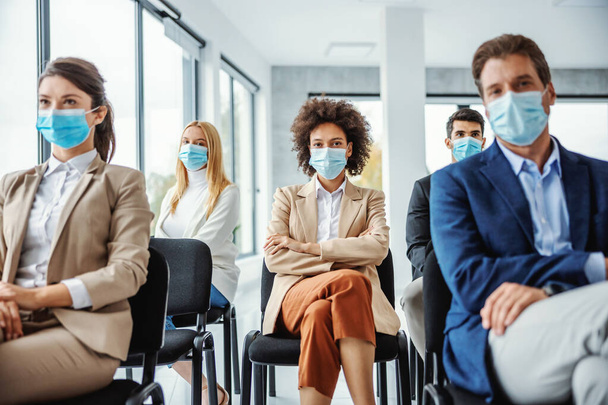 コロナウイルスのセミナーに座って顔マスクを持つビジネスマンの多人種グループ。途中で混合人種の女性に選択的な焦点. - 写真・画像