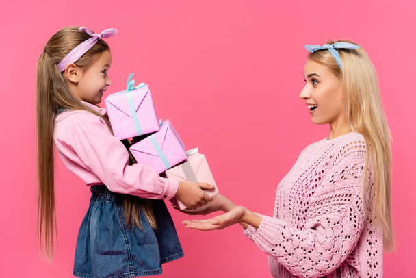 вид изумленной матери, смотрящей на дочь с подарками, изолированными на розовом фоне - Фото, изображение