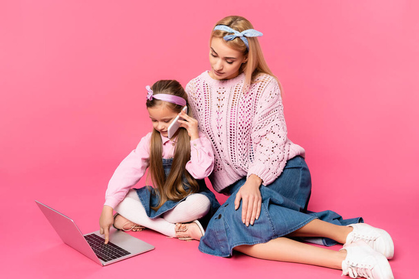 kid talking on smartphone and using laptop while sitting with mother on pink - Foto, Imagen