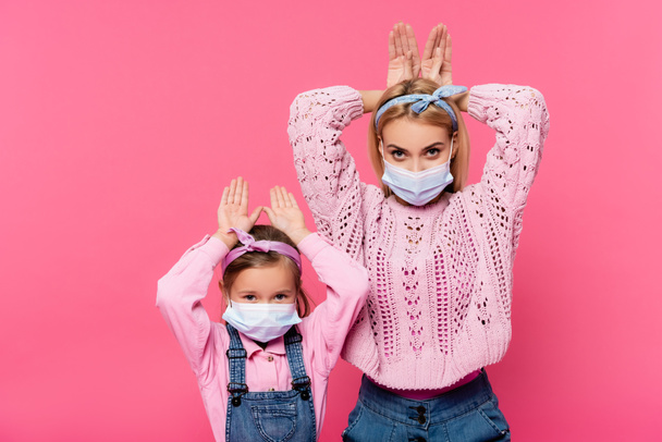 mère et fille dans des masques médicaux faisant des oreilles de lapin avec les mains isolées sur rose  - Photo, image