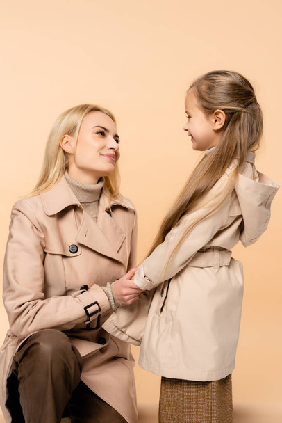 mother and daughter in trench coats holding hands while looking at each other isolated on beige - Photo, Image
