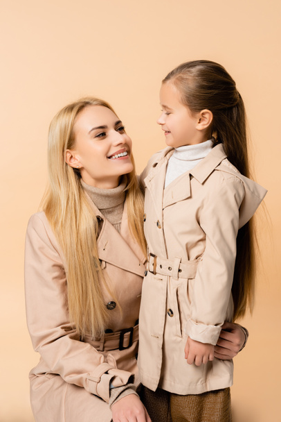 joyful mother and daughter in trench coats looking at each other and smiling isolated on beige - Foto, Bild