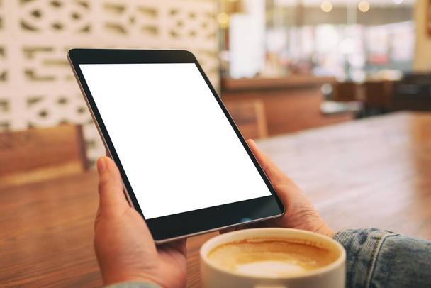 Mockup afbeelding van handen met zwarte tablet pc met blanco wit scherm met koffiekopje op houten tafel  - Foto, afbeelding