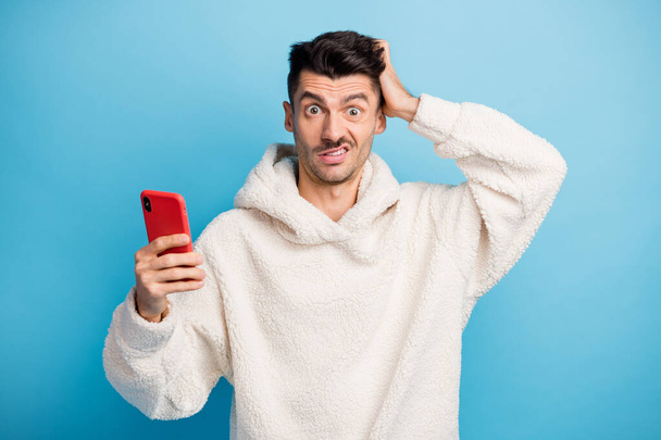 Photo portrait of man scratching head holding phone in one hand wearing wool hoodie isolated on pastel blue colored background - 写真・画像