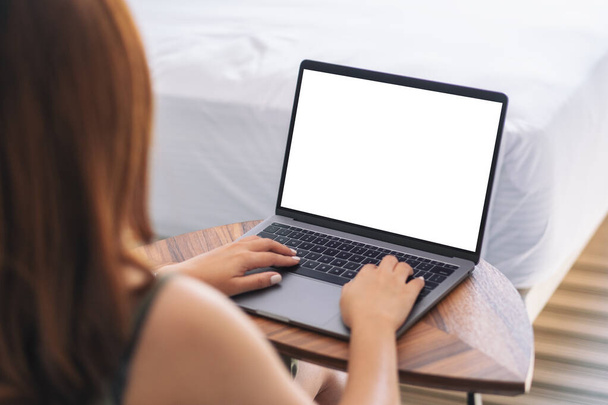 Mockup beeld van een vrouw met en typen op laptop met blank wit bureaublad toetsenbord op houten tafel naast het bed - Foto, afbeelding