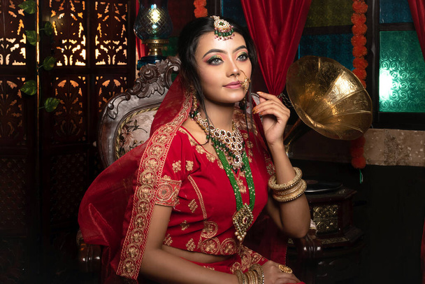 Magnífico jovem noiva indiana em vestido luxuoso e jóias preciosas está sentado em uma cadeira em um apartamento de luxo. Interior vintage clássico. Moda de casamento. - Foto, Imagem