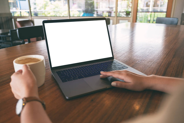 Mockup image of a woman using and touching on laptop touchpad with blank white desktop screen on wooden table while drinking coffee - Foto, Imagem