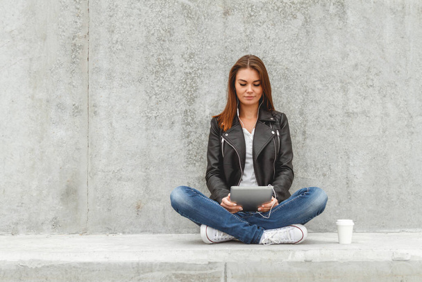 Girl with a tablet and headphones in hands sits on a concrete wall. Online education students - Foto, imagen
