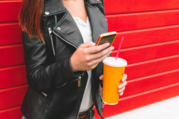 lány mobiltelefont használ, SMS-t ír, egy itallal a kezében egy piros fa falon - Fotó, kép