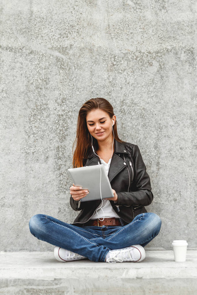 Girl with a tablet and headphones in hands sits on a concrete wall. Online education students - Fotoğraf, Görsel