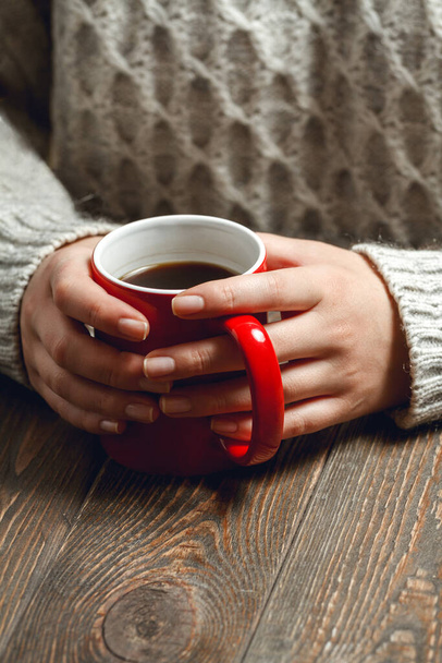 The girl in a cozy knitted sweater drinking coffee from a red mug, sitting at a vintage wooden table - Foto, imagen