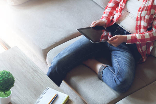 Woman sitting on sofa uses tablet, holding in hands - Fotoğraf, Görsel