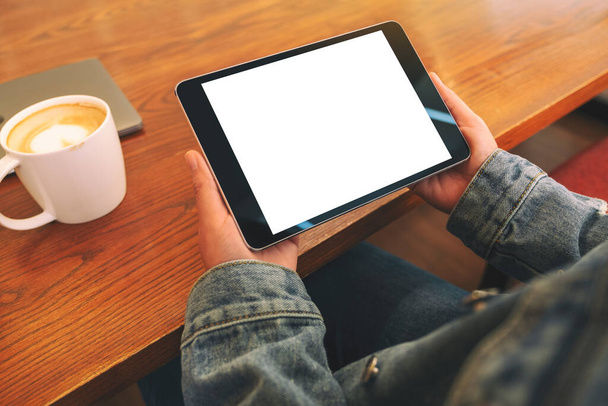 Imagem Mockup de mãos segurando tablet preto pc com tela branca em branco horizontalmente com xícara de café na mesa de madeira  - Foto, Imagem