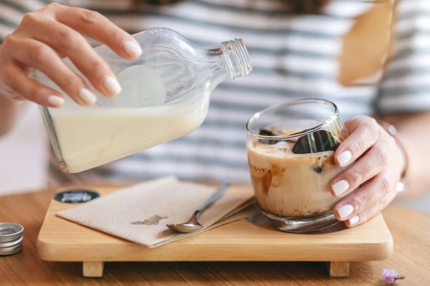 Imagen de cerca de una mujer vertiendo leche en un vaso de café con hielo en la cafetería - Foto, imagen