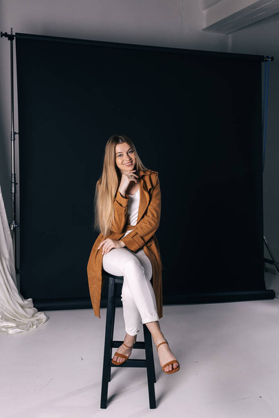 Muodikas nainen tyylikkäissä vaatteissa poseeraa tummalla taustalla studiossa. Kaunis tyttö yllään ruskea takki, valkoiset housut, istuu tuolilla. - Valokuva, kuva