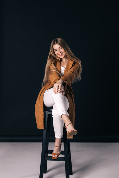 Modieuze vrouw in stijlvolle kleding poseren op donkere achtergrond in de studio. Mooi meisje in bruine jas, witte broek, zittend op stoel. - Foto, afbeelding