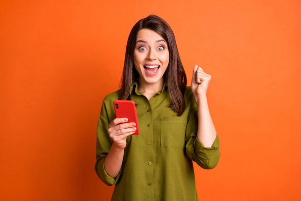 Photo of crazy funky girl hold cellphone raise fist open mouth wear green shirt isolated orange color background - Foto, afbeelding