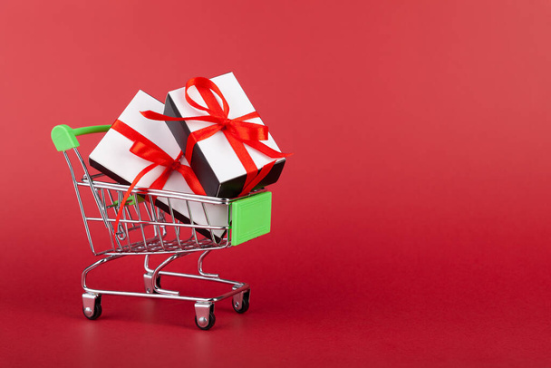 Caja de regalo con cinta roja en mini carrito de comestibles sobre fondo rosa - Foto, Imagen