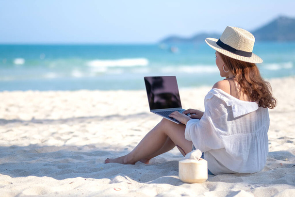 Eine Frau tippt auf der Tastatur eines Laptops, während sie an einem schönen Strand sitzt - Foto, Bild
