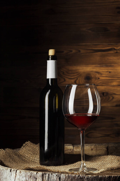 Bottle and glass of red wine on wooden barrel shot with dark wooden background - Фото, изображение