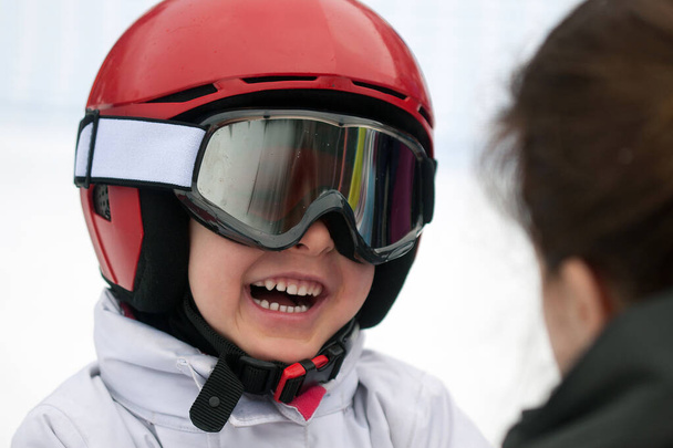 Cheerful boy in red helmet, ski goggles and white jacket smiling to his mother. Winter sports, young skier, snowy background - Photo, Image